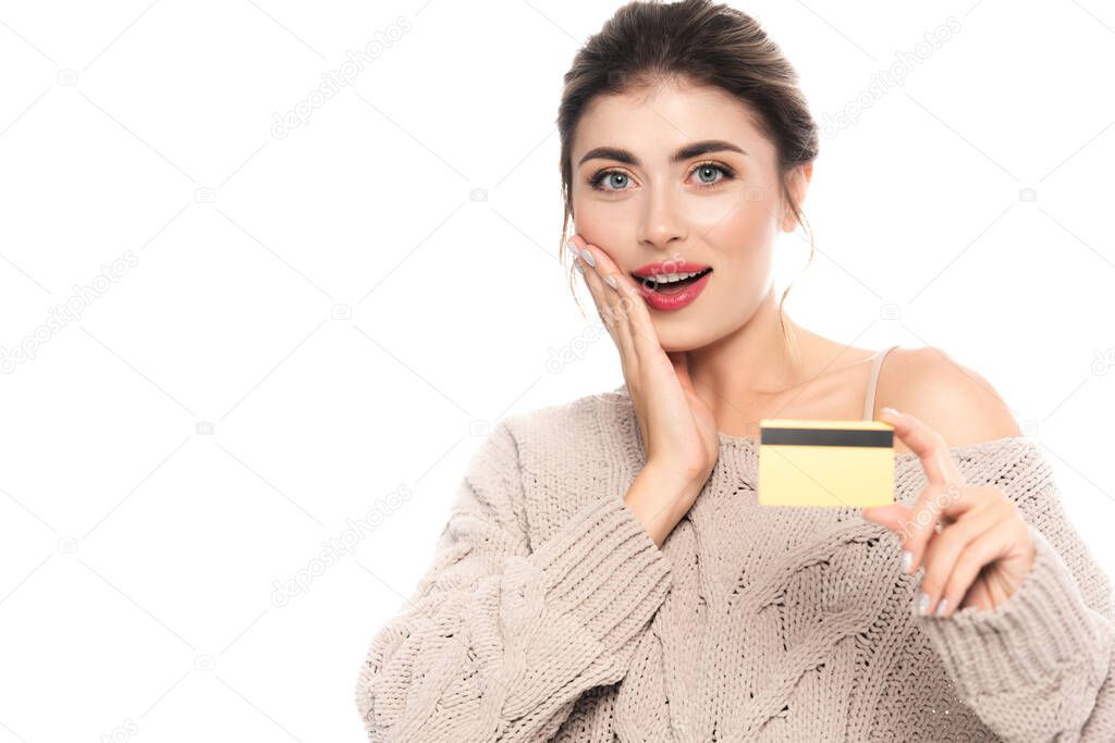 excited woman in trendy sweater showing credit card while looking at camera isolated on white