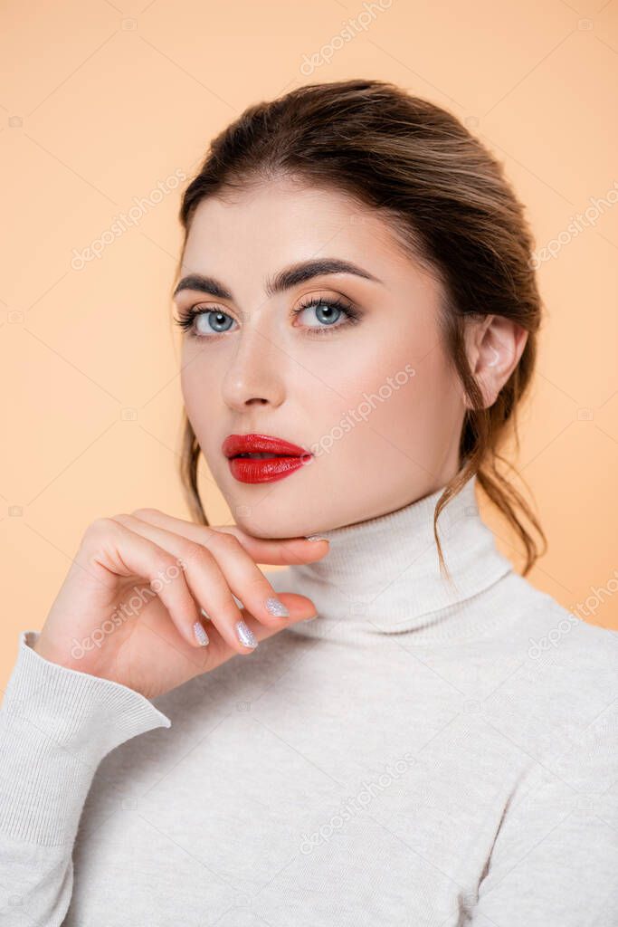 stylish woman in turtleneck holding hand near chin while looking at camera isolated on peach 