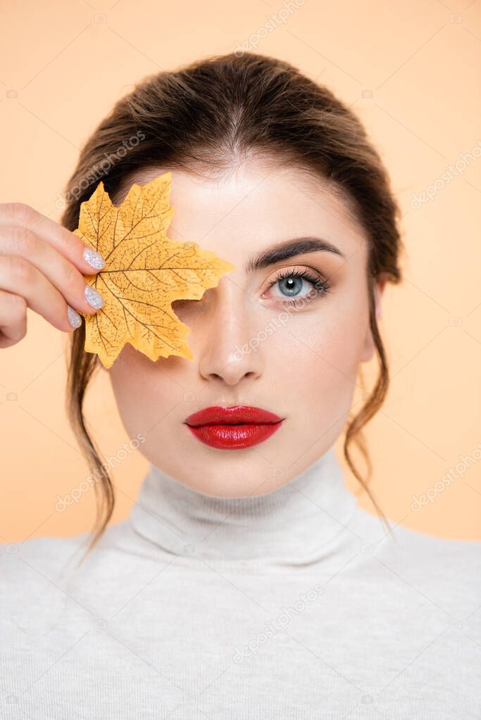 young woman with red lips covering eye with yellow leaf while looking at camera isolated on peach 