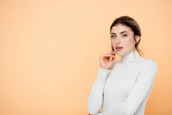 stylish woman in turtleneck holding hand near face isolated on peach 
