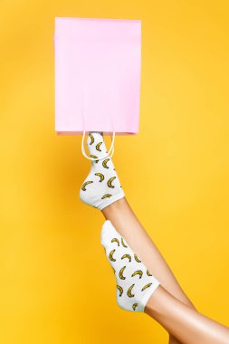 Çorap giyen bir kadın, sarı arka planda izole edilmiş pembe bir kesekağıdına bacağını koyuyor.
