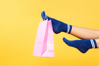 Sarı arka planda izole edilmiş çoraplarla kadın bacağına asılı pembe kağıt torba görüntüsü.