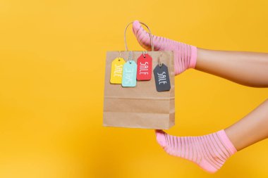 Cropped view of paper bag with sale tags hanging on woman legs in socks isolated on yellow  clipart