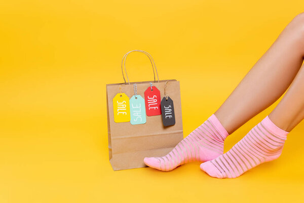 Cropped view of female legs in socks with pink stripes near paper bag with tags with sale words isolated on yellow