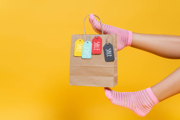 Cropped view of paper bag with sale tags hanging on woman legs in socks isolated on yellow 
