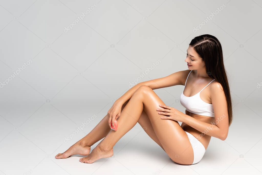 Brunette woman in white underwear, shaving leg with pink epilator, while sitting on grey