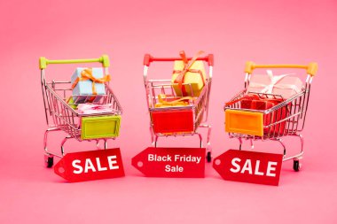 colorful presents in shopping trolleys with sale tags on pink, black friday concept  clipart