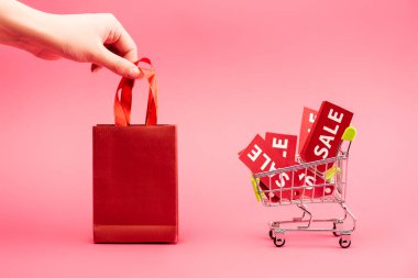cropped view of female hand touching shopping bag near cart with sale labels on pink clipart