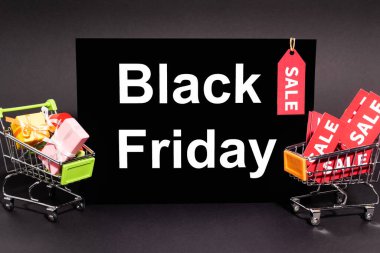 small presents in toy shopping carts near placard with black friday lettering and sale tag on dark background  clipart