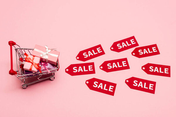 top view of toy shopping cart with presents near tags with sale lettering on pink, black friday concept 