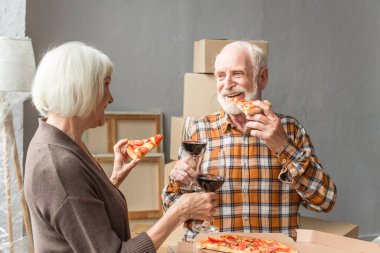 Yeni evde pizza yiyen ve şarap bardakları tutan gülen yaşlı çift.