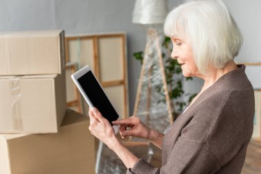 Arka planda karton kutular, hareketli konsept ile boş ekranlı dijital tablet tutan yaşlı kadının yan görünümü