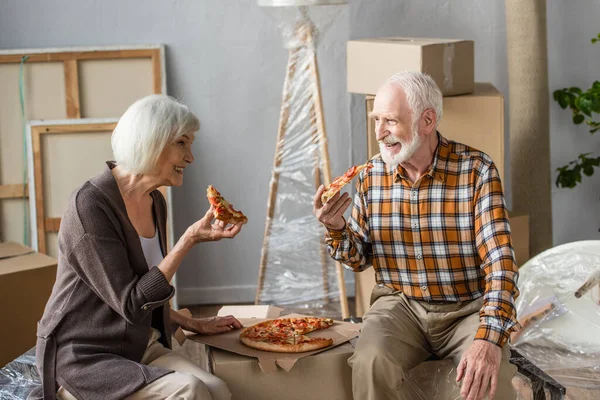 laughing senior couple eating pizza in new house and cardboard boxes on background