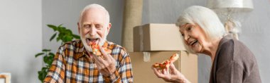 Yeni evde pizza yiyip arka planda karton kutular yiyen yaşlı çiftin panoramik görüntüsü.