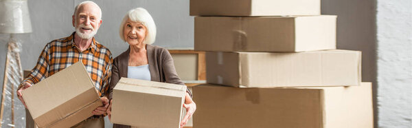 panoramic shot of senior couple holding cardboard box and looking at camera in new house, moving concept