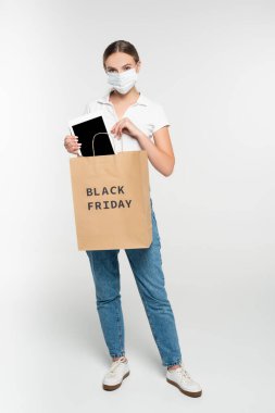 Tıbbi maskeli ve kot pantolonlu bir kadın, beyaz harflerle siyah cuma harfleriyle dolu alışveriş çantasına boş ekranlı dijital tablet koyuyor. 