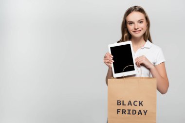 Neşe dolu genç bir kadın elinde gri harflerle alışveriş çantasının yanında boş ekranlı dijital tablet tutuyor.