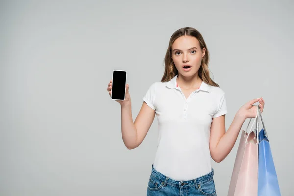 Geschokte Vrouw Met Smartphone Met Leeg Scherm Boodschappentassen Geïsoleerd Grijs — Stockfoto