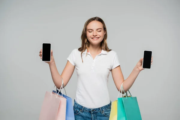 joyful woman with closed eyes holding mobile phones with blank screen and shopping bags isolated on grey, black friday concept