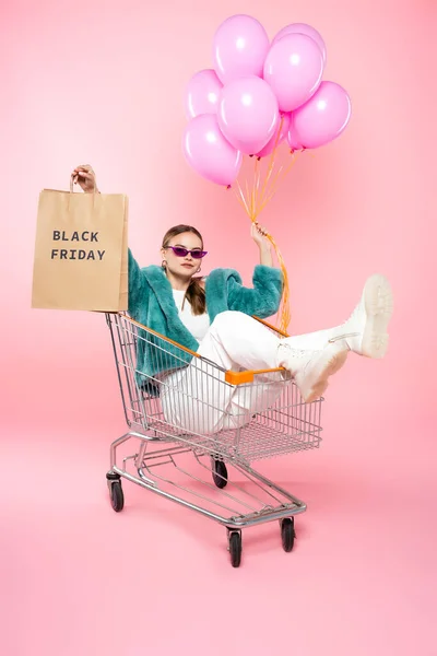 戴着太阳镜的时髦女人坐在购物袋上印有黑色星期五字母的购物车里 手里拿着粉色气球 — 图库照片