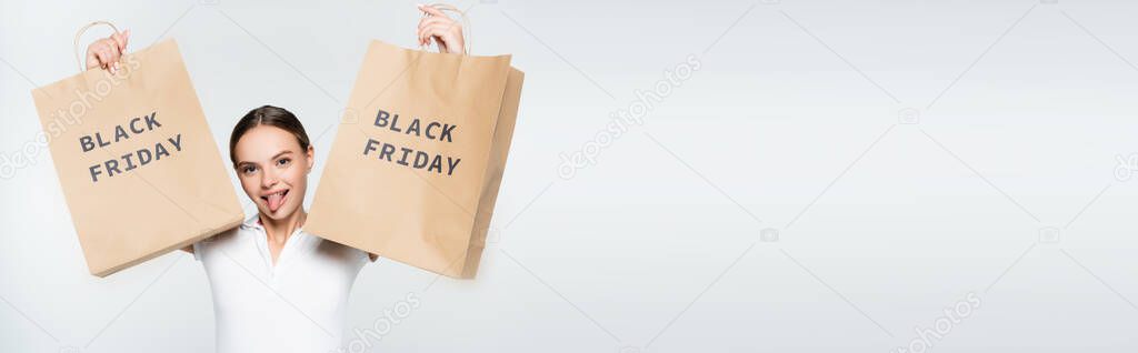 panoramic shot of woman sticking out tongue while holding shopping bags with black friday lettering on white