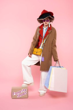 Güneş gözlüklü ve şapkalı bir kadın elinde alışveriş çantaları ve yanında pembe harflerle en sevdiği ayakkabılarla duruyor.