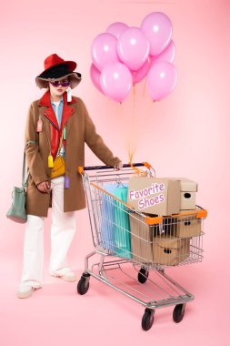 Güneş gözlüklü ve şapkalı moda bir kadın alışveriş çantaları ve pembe harfli kutularla alışveriş arabasının yanında duruyor.