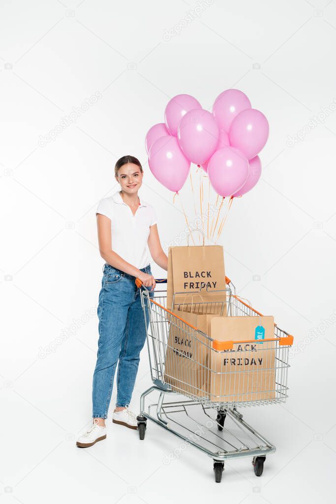 pleased woman standing near cart with shopping bags, black friday lettering and pink balloons on white