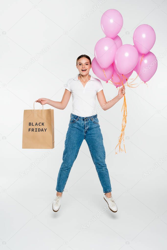 joyful woman holding shopping bag with black friday lettering and pink balloons while jumping on white