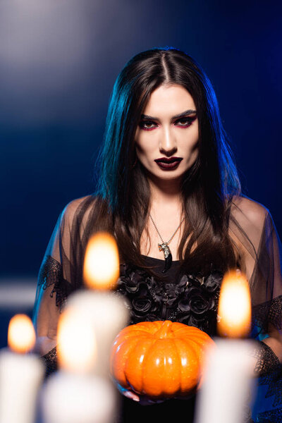 selective focus of young woman with black makeup holding pumpkin near burning candles on blue, halloween concept