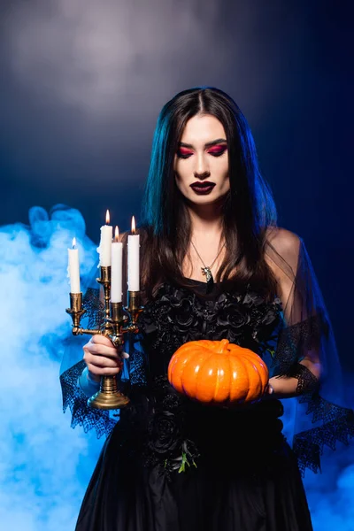 pale woman with black makeup holding pumpkin and burning candles on blue with smoke, halloween concept