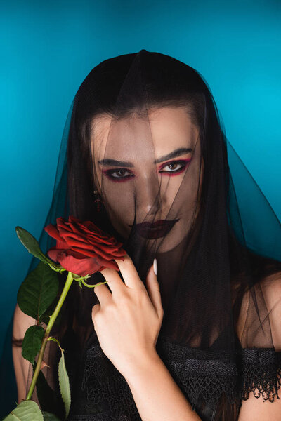 evil woman with black makeup looking at camera through veil near rose on blue