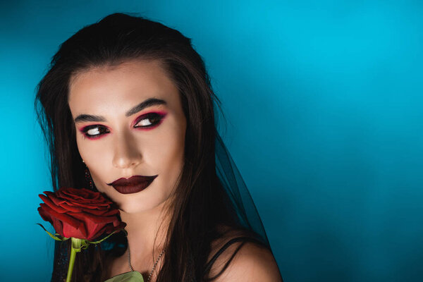 young woman with black makeup in veil looking away near rose on blue