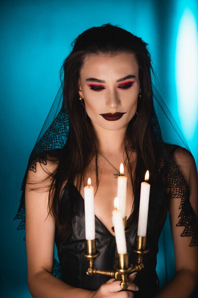 selective focus of woman with black makeup and veil looking at burning candles on blue