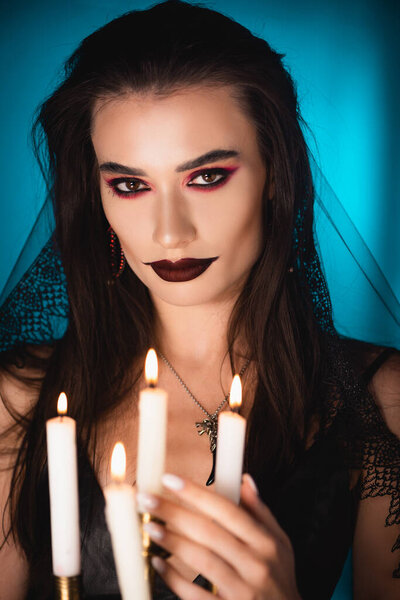 selective focus of young woman with black makeup and veil near burning candles on blue