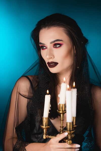 pale woman with black makeup looking away and holding burning candles on blue