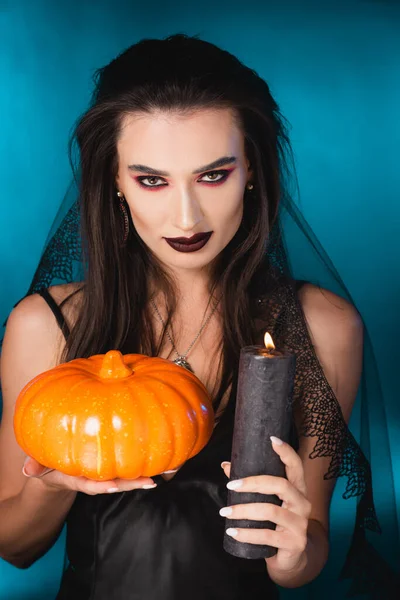 pale woman with black makeup and veil holding burning candle and pumpkin on blue