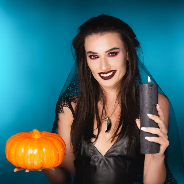 young pale woman with makeup and veil holding burning candle and pumpkin on blue