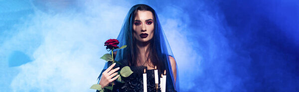 panoramic crop of woman in black veil holding rose and burning candles on blue with smoke, halloween concept