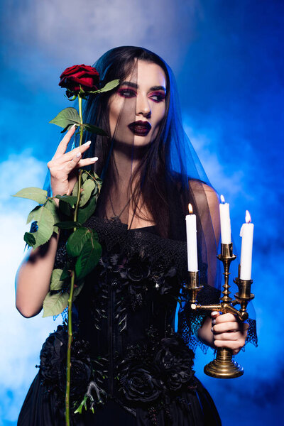 young woman in black dress and veil holding rose and burning candles on blue with smoke, halloween concept