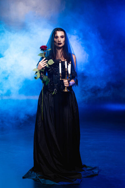 bride in black dress and veil holding  rose and burning candles on blue with smoke, halloween concept