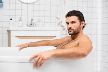Sakallı adam evde banyo yaparken kameraya bakıyor. 