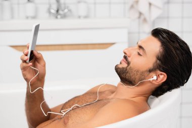 Kulaklıklı genç adam banyo yapıyor ve akıllı telefon kullanıyor. 