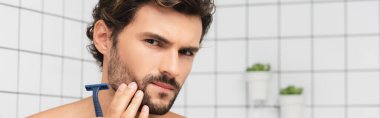 Man looking at camera while holding razor in bathroom, banner  clipart