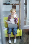 pohled přes sklo blondýny v kostkované košili pracovní z domova a pití čaje