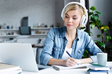 Kulaklıklı genç sarışın kadın internette ders çalışıyor ve notlar alıyor.