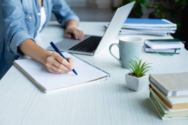 cropped view of woman making notes in notebook and typing on laptop clipart