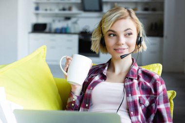young blonde woman in headphones working from home and drinking tea clipart