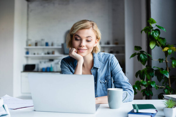 young blonde woman leaning chin on hand and working on laptop
