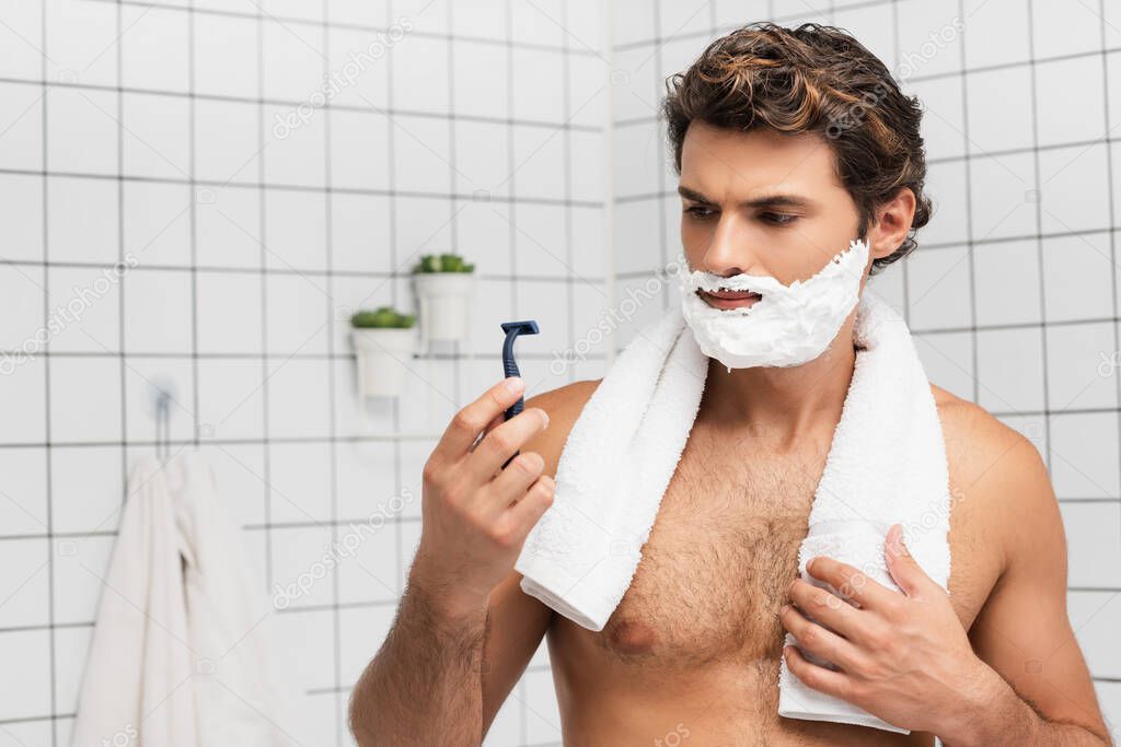 Man with shaving foam on face holding razor and towel in bathroom 